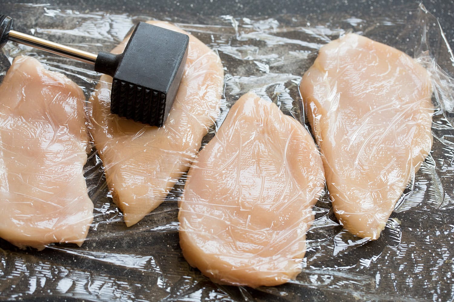 Flattening chicken breasts with a meat mallet on a cutting board.