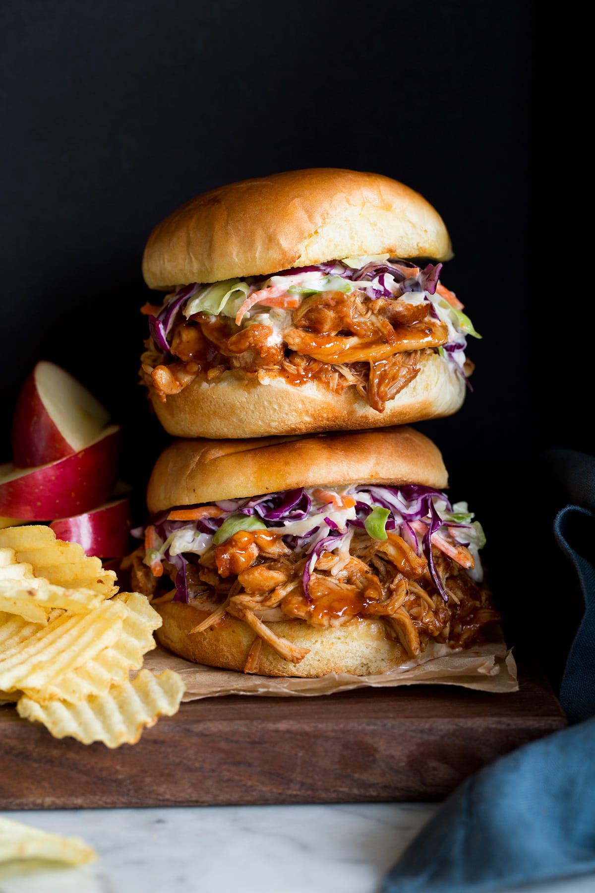 Crockpot BBQ Chicken Photo: Two BBQ Pulled Chicken Sandwiches stacked on a wood platter. Apples slices and potato chips are shown to the side as serving suggestions.