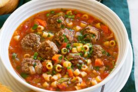 Serving of meatball soup in a white bowl over a white plate with bread to the side.