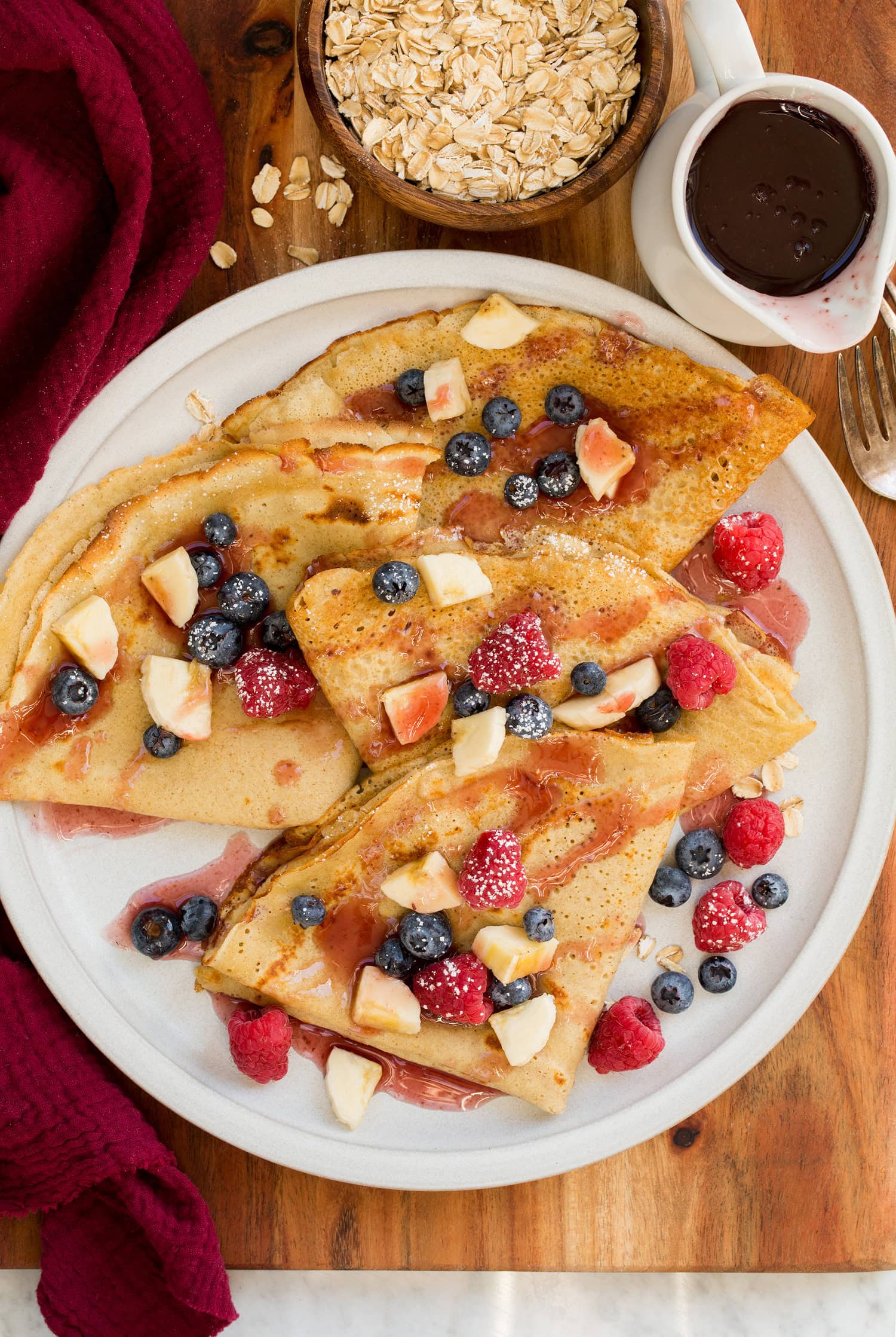 Overhead photo of oat crepes on w white plate set on a wood board with a red cloth to the side along with syrup and oats.