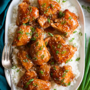 Slow cooker chicken thighs with sauce shown served over white rice on a white oval platter. Platter is on a marble surface with a blue cloth and green onions to the side.