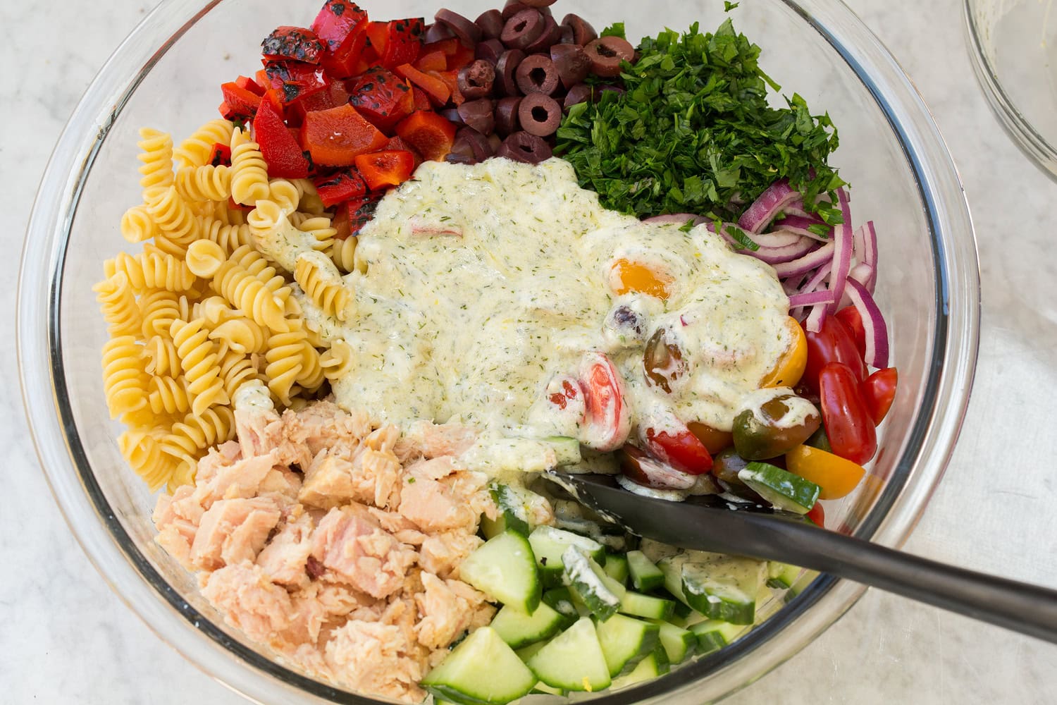 Pasta salad ingredients including tuna, vegetables, olives and creamy dressing in a bowl shown before mixing.